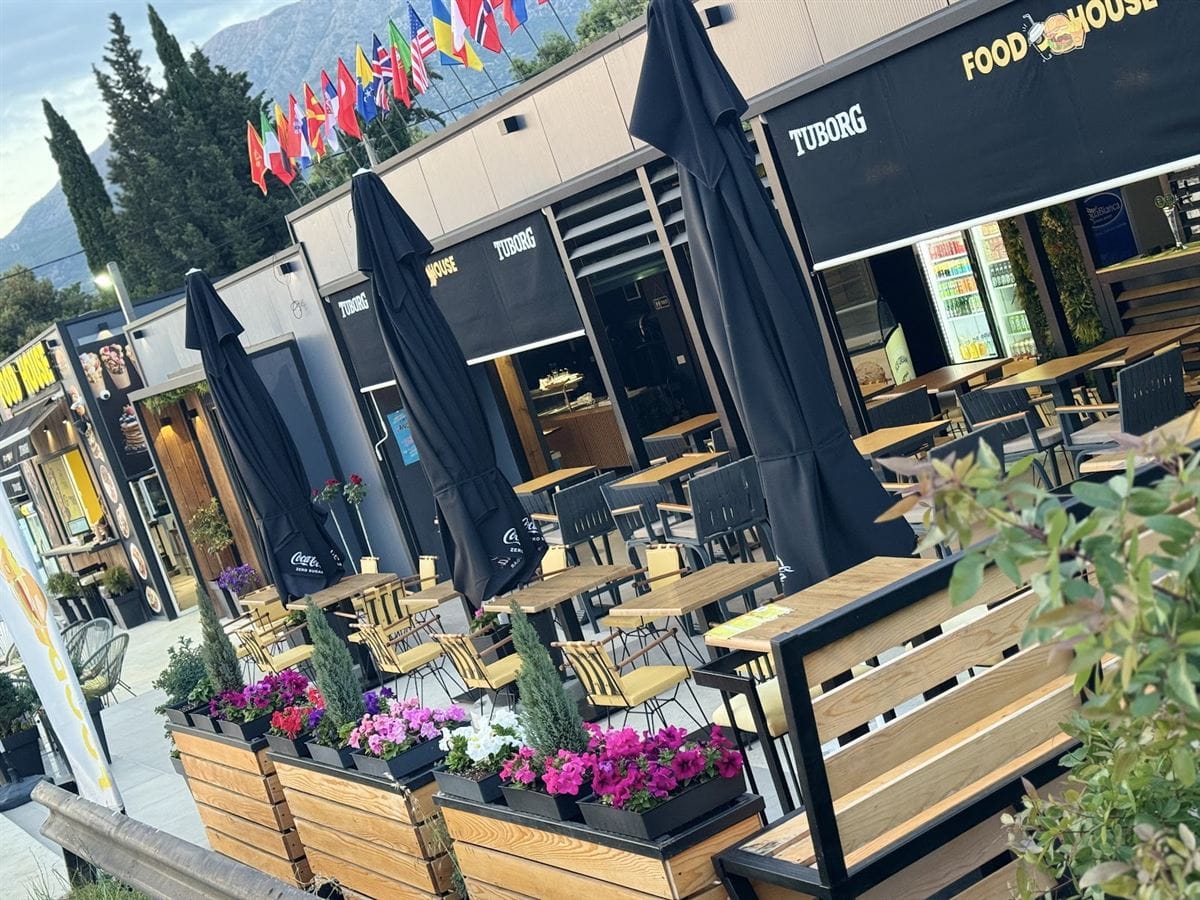 Die überdachte Terrasse in der Dämmerung: Holztische, Blumenkästen und eine Reihe von Länderflaggen.